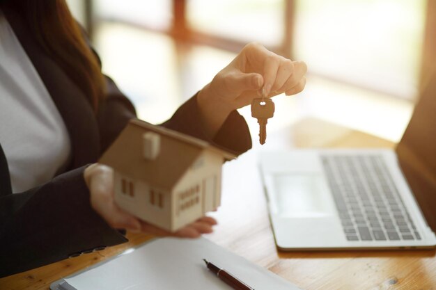 Une femme courtier immobilier tenant un porte-clés à la maison et un modèle de maison dans sa main est assise au bureau au bureau