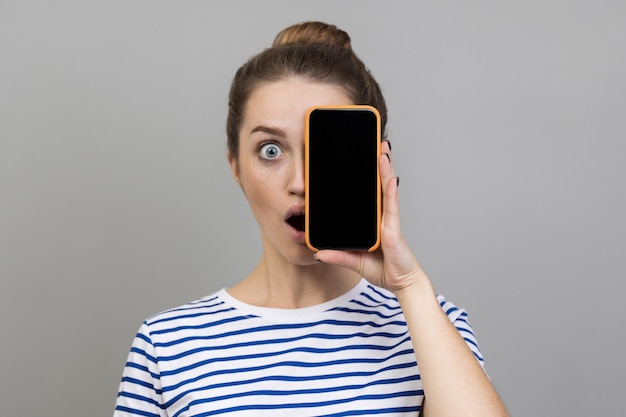 Femme couvrant la moitié du visage avec un téléphone portable et regardant avec surprise la technologie et l'identité de l'appareil photo