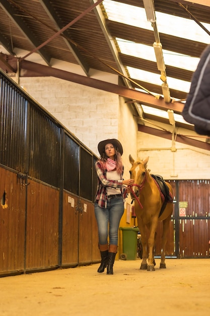 Femme de cow-girl caucasienne marchant à cheval dans une écurie, chapeaux du sud des États-Unis, chemise à carreaux rose et jeans