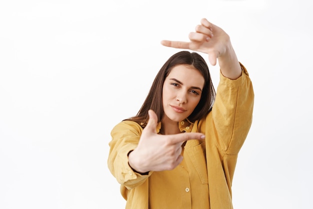 Femme créative réfléchie à la recherche d'un coup parfait faisant des cadres de main geste de la caméra et regardant à travers un chef-d'œuvre de création pensif debout sur fond blanc