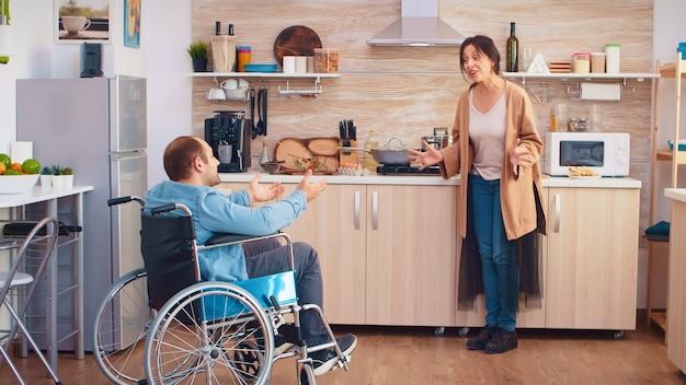 Femme criant dans la cuisine à son homme handicapé en fauteuil roulant tout en ayant un désaccord. Guy avec paralysie handicap handicap handicapé difficultés à obtenir de l'aide pour la mobilité de l'amour et de la relation