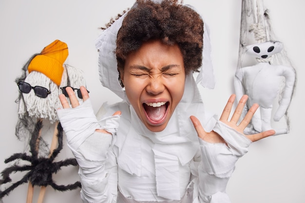 La femme crie fort garde la bouche largement ouverte les mains levées vêtues d'un costume fantôme isolé sur blanc avec des jouets effrayants faits à la main qui traînent