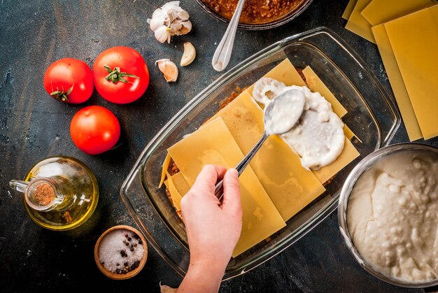 Femme cuisinant des lasagnes bolognaises classiques faites maison, sur une table bleu foncé; avec des ingrédients, vue de dessus copie espace, mains en photo