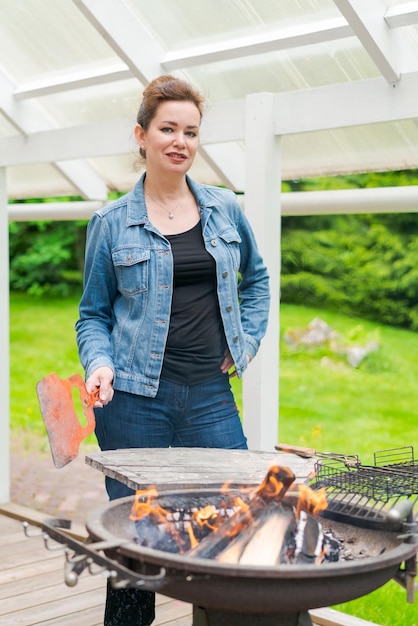 Une femme cuisine un barbecue pour sa famille à l'extérieur dans le jardin d'une maison de campagne portant