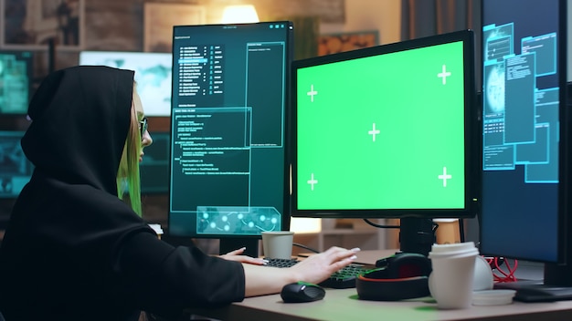 Femme cyber terroriste portant un sweat à capuche travaillant sur ordinateur avec écran vert.