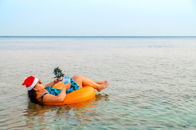 Une femme dans un anneau de natation dans un chapeau de père Noël avec un cocktail et un petit arbre de Noël à la main près du rivage dans la mer. Tir de dos. photo horizontale