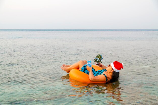 Une femme dans un anneau de natation dans un chapeau de père Noël avec un cocktail et un petit arbre de Noël à la main près du rivage dans la mer. Tir de dos.