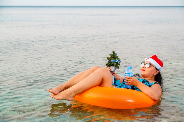 Une femme dans un anneau de natation dans un chapeau de père Noël avec un cocktail et un petit arbre de Noël