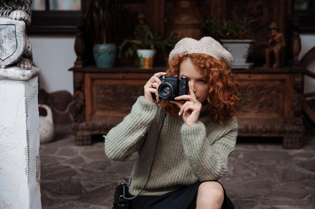 Une femme dans un béret et un pull vert tient un appareil photo dans ses mains et prend des photos