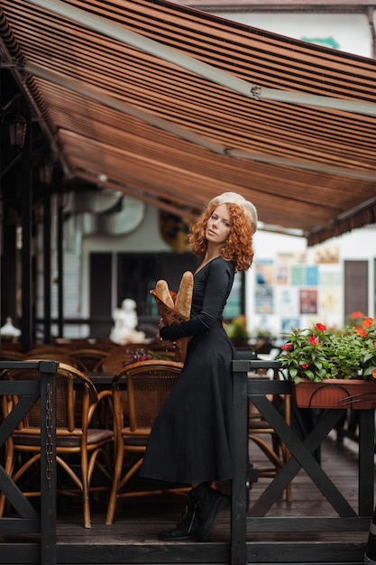 Une femme dans un béret et une robe noire tient une baguette dans ses mains du fond c