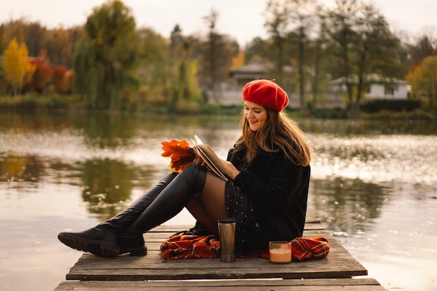 Femme dans un béret rouge lisant un livre sur un ponton en bois Saison d'automne Femme avec des vêtements à la mode se détendre près de l'eau lisant un livre et buvant du café Ambiance d'automne Concept d'automne