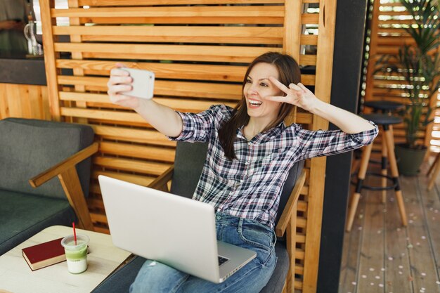 Femme dans un café d'été en bois à l'extérieur de la rue assise avec un ordinateur portable, faisant une photo de selfie sur un téléphone portable, se relaxant pendant le temps libre. Bureau mobile