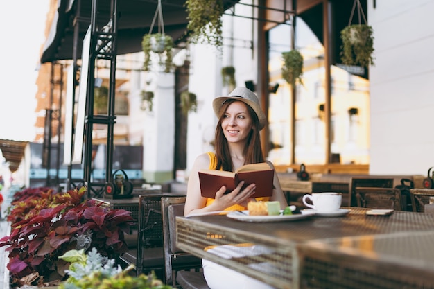 Femme dans un café de rue en plein air assis à table dans un chapeau, lisant un livre avec une tasse de cappuccino, un gâteau, se relaxant au restaurant pendant le temps libre