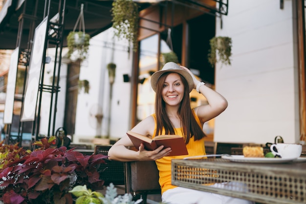 Femme dans un café de rue en plein air assis à table dans un chapeau, lisant un livre avec une tasse de cappuccino, un gâteau, se relaxant au restaurant pendant le temps libre