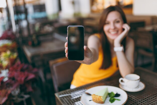 Femme dans un café de rue en plein air assis à table avec une tasse de thé, un gâteau, tenir dans la main un téléphone portable avec un écran vide vierge