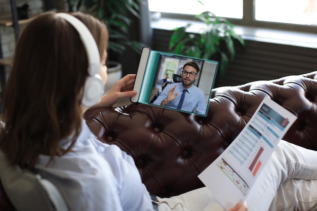 Une femme dans un casque allongé sur un canapé parle par appel vidéo avec des collègues lors d'un briefing en ligne sur une tablette numérique Pavé tactile moderne transparent du futur concept