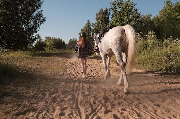 Femme dans un casque marche avec son cheval le long d'une route sablonneuse poussiéreuse en la tenant par la bride