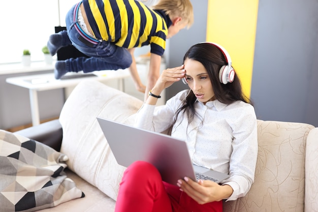 Femme dans un casque et un ordinateur portable est assis sur un canapé à côté de chouchouter enfant