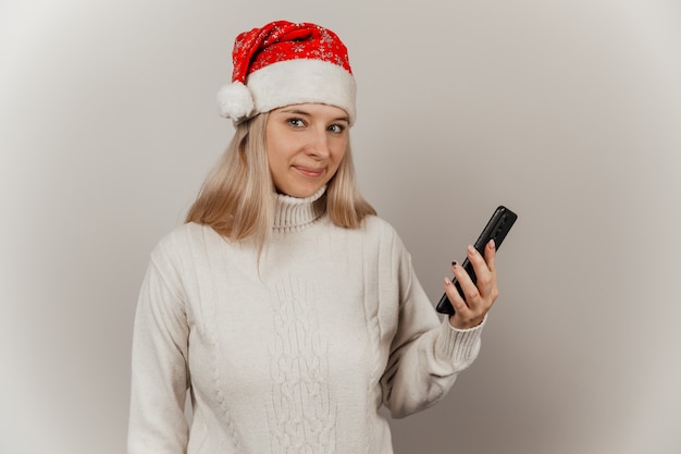 Femme dans un chandail blanc et un bonnet de Noel avec un téléphone dans ses mains sur un fond gris isolé