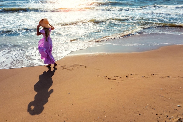 Femme dans un chapeau de paille avec les mains en l'air marche sur la plage