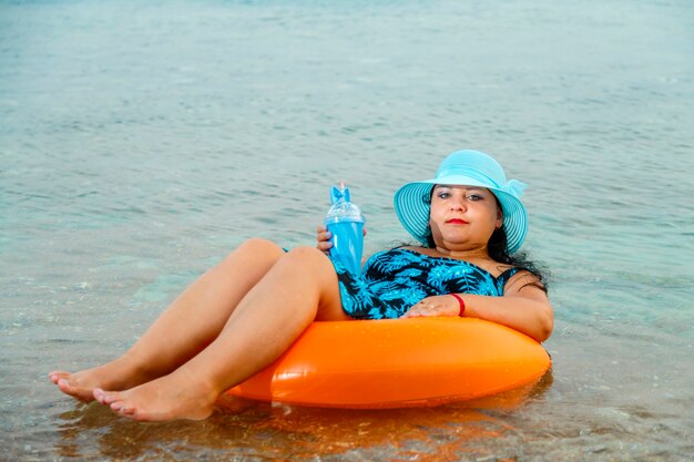 Une femme dans un chapeau de paille et des verres dans la mer dans un cercle de natation avec un cocktail à la main