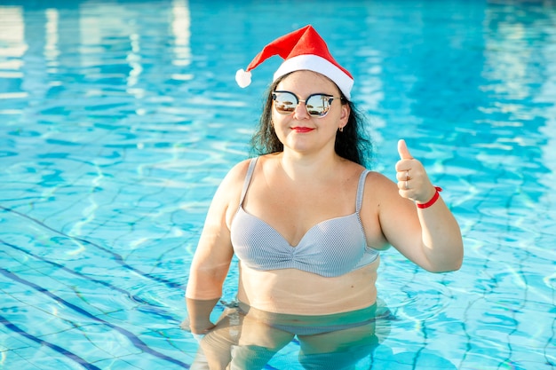 Une femme dans un chapeau de Père Noël et des lunettes de soleil dans la piscine célèbre Noël et montre sa classe