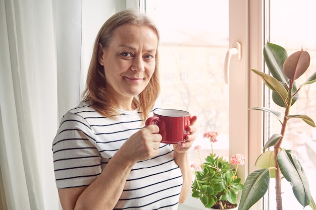 femme dans une chemise rayée boit du thé en se tenant debout près de la fenêtre