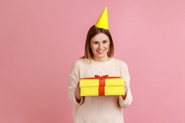 Femme dans un cône de fête tenant une boîte-cadeau emballée jaune regardant la caméra avec une expression satisfaite