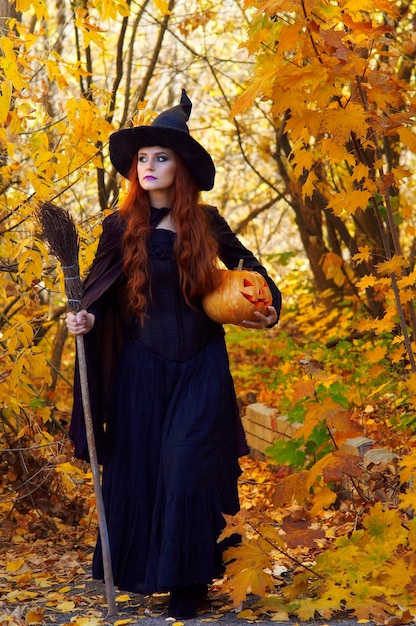 Une femme dans un costume de sorcière noire et un chapeau pointu avec un balai et une citrouille dans un parc à l'Halloween