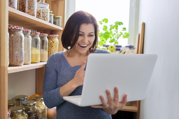 Femme dans le garde-manger de la cuisine avec des produits stockés tenant un ordinateur portable