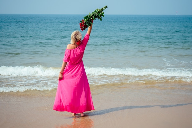 Une femme dans une longue robe rose bénéficie d'un bouquet de roses sur la plage