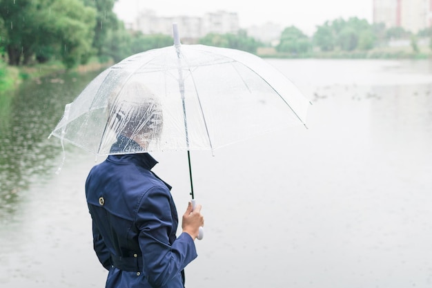 Une femme dans un manteau bleu est tournée avec son dos sous un parapluie transparent à l'automne près du lac