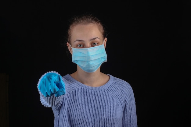 Femme dans un masque médical et un gant vous montre sur un mur noir.