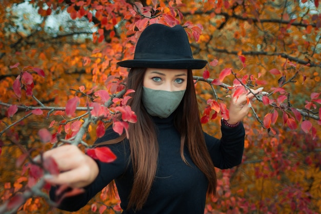 Une femme dans un masque de protection est vêtue d'une veste noire et d'un chapeau contre un parc d'automne