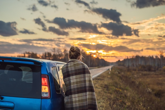 Une femme dans un plaid se tient près de la voiture sur le bord de la route à l'arrière-plan de l'aube Concept de voyage en voiture