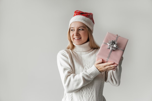 Femme dans un pull blanc et un bonnet de Noel rouge tenant un cadeau rose dans ses mains sur fond gris