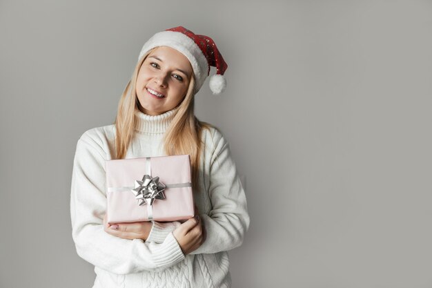 Femme dans un pull blanc et un bonnet de Noel rouge tenant un cadeau rose dans ses mains sur fond gris