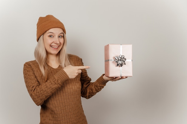 Femme dans un pull marron et un chapeau tenant un cadeau rose dans ses mains sur fond gris