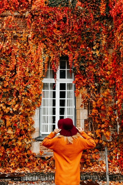 Femme dans un pull orange et un chapeau près d'un mur avec des raisins sauvages en automne