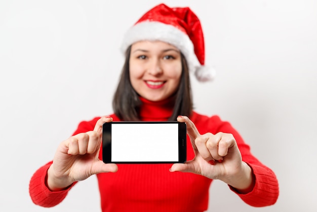 Femme dans un pull rouge et un chapeau de Noël montre un smartphone avec un écran blanc vierge