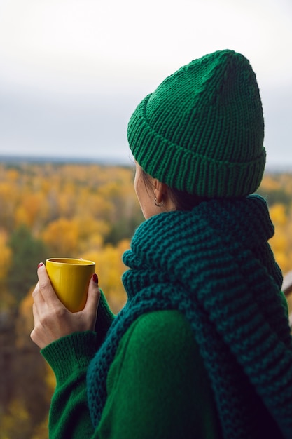 Femme dans un pull en tricot vert et un chapeau se dresse avec une tasse jaune sur le balcon dans le contexte d'une forêt d'automne
