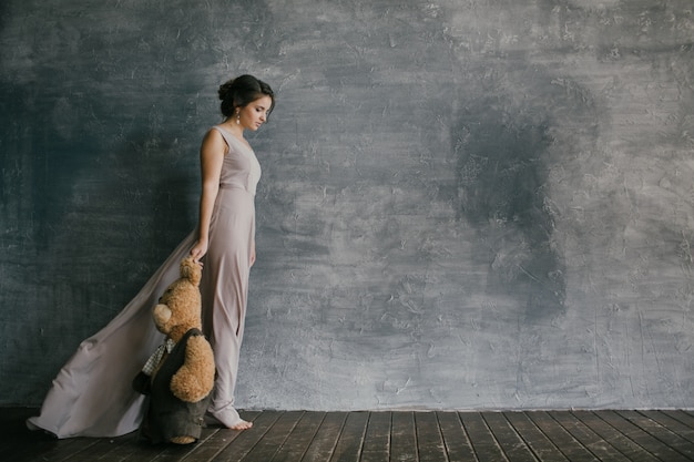 femme dans une robe rose marche à côté d'un mur gris et détient un ours en peluche