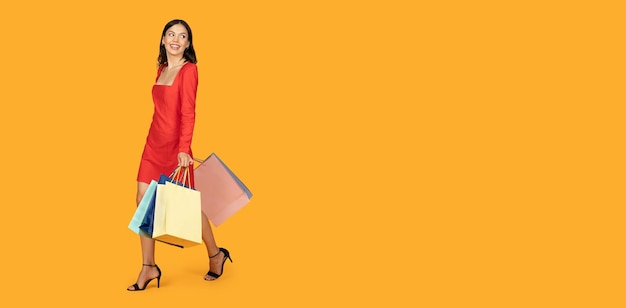 Une femme dans une robe rouge marche vers le côté droit du cadre portant plusieurs sacs d'achat.