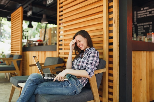 Femme dans la rue en plein air café d'été café en bois assis dans des vêtements décontractés, travaillant sur un ordinateur portable moderne