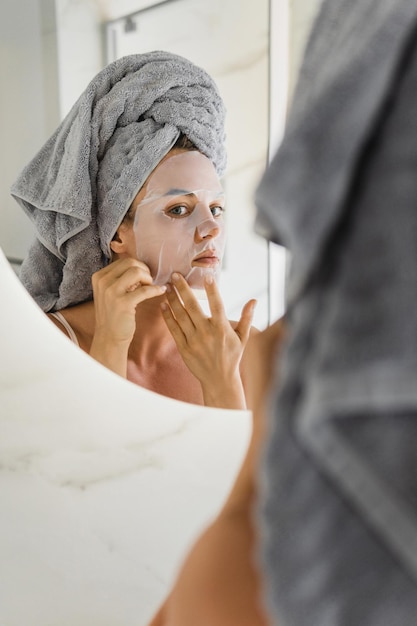 Femme Dans La Salle De Bain Avec Masque En Feuille Appliqué Sur Son Visage Regardant Dans Le Miroir