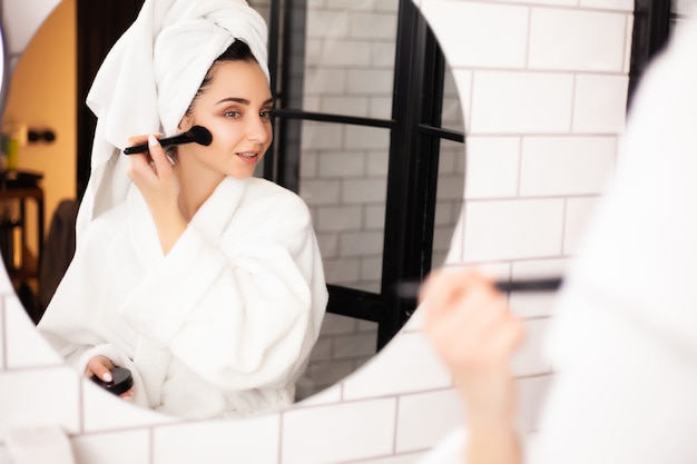 Une femme dans la salle de bain avec une serviette sur la tête applique du maquillage sur son visage.