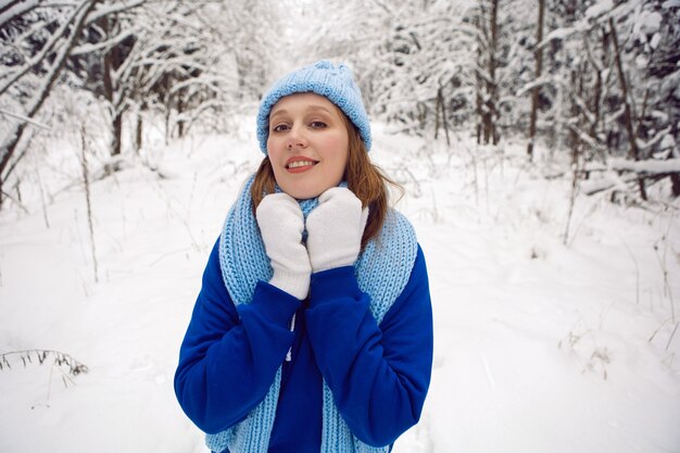 Femme dans un survêtement bleu mitaines blanches et écharpe se dresse en hiver dans une forêt couverte de neige