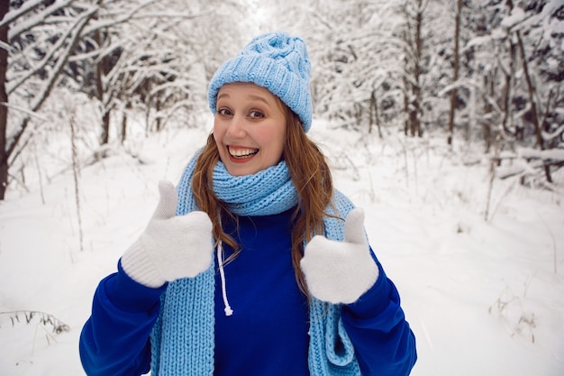 Femme dans un survêtement bleu mitaines blanches et écharpe se tient les pouces vers le haut en hiver dans une forêt couverte de neige