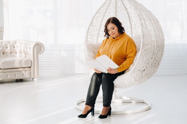 Une femme dans un sweat à capuche orange est assise sur une chaise inhabituelle avec un livre dans ses mains.