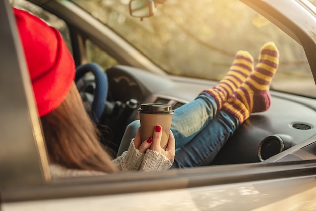 Femme dans une voiture en chaussettes de laine chaude jaune tient une tasse de café dans les mains au coucher du soleil